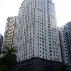 The AIC Gold Tower is a white building with a brown podium facade and several rectangular and semi-oblong recesses. Several adjacent buildings are partly visible on its side.