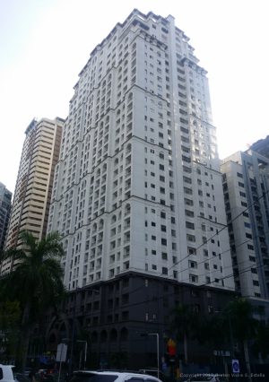 The AIC Gold Tower is a white building with a brown podium facade and several rectangular and semi-oblong recesses. Several adjacent buildings are partly visible on its side.
