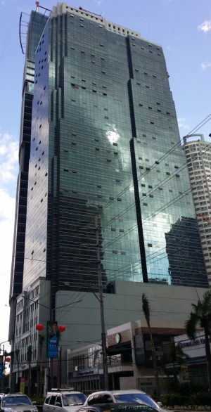 Antel Global Corporate Center is a tall cuboid structure with a lower podium of a teal concrete exterior at about one-fourth of its height and upper levels of a teal glass facade. This is the building as seen from Garnet Road corner Julia Vargas Ave.
