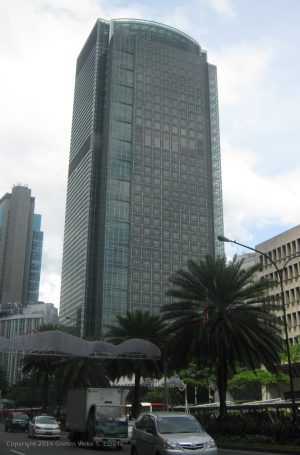 Ayala Tower One is a skyscraper with facade made of square glass panels reflecting a cloudy sky, surrounded by palm trees and a busy street. This is the tower as seen from sidewalk in front of Allied Bank Center.