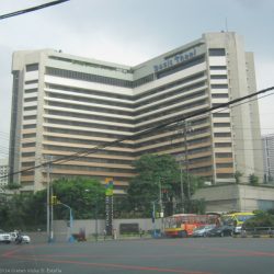 The facade of Dusit Thani Manila features a pattern of horizontal stripes of concrete walls and indented glass panels. It is surrounded by a concrete barrier that has greenery growing within it. This is the hotel as seen from opposite corner on EDSA northbound side.