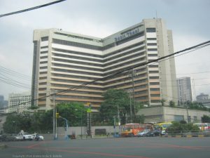 The facade of Dusit Thani Manila features a pattern of horizontal stripes of concrete walls and indented glass panels. It is surrounded by a concrete barrier that has greenery growing within it. This is the hotel as seen from opposite corner on EDSA northbound side.