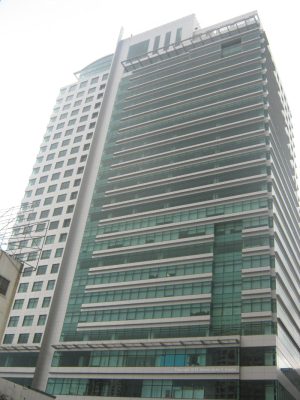 Gateway Tower is a gray rectangular prism with a concrete and glass facade and uneven divide. The larger part has bands of gray concrete and green glass panels, while the smaller part has small square windows. The photo was taken from the sidewalk along Gen. Malvar Ave. near Cambridge St.