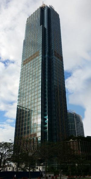 Metrobank Center is a tall rectangular prism covered with dark green glass. The top of the structure, which is a beige truncated pyramid, is barely visible from this shot taken from within the vacant lot surrounded by 35th St., 9th Ave., 34th St., and 8th Ave.