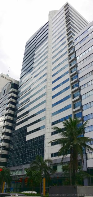 Hanston Square has a blocky structure and an exterior of dark glass and white concrete. The view from the sidewalk on the opposite side of San Miguel Ave. and adjacent to Lourdes School is shown here.