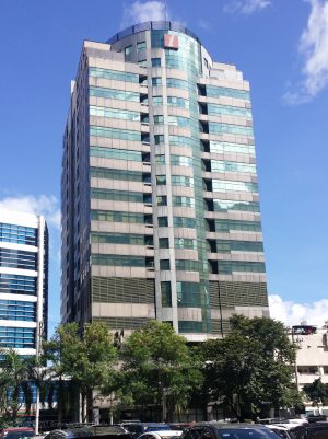 I-Square Building is a tall cuboid with a vertical semi-cylindrical protrusion in front. The upper floors have gray concrete and blue glass facade, while the lower floors have steel-covered windows. This is I-Square, as seen from the parking between Sapphire Road and Garnet Road near Julia Vargas Avenue.