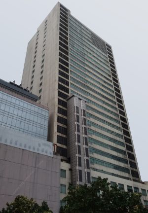 IBM Plaza has a tall blocky appearance and has a facade of mostly gray concrete. It also has what looks like an external elevator shaft that reaches halfway to the floors above the podium. This picture shows the back of IBM Plaza, as seen from the compound of St. Pio of Pietrelcina Chapel.