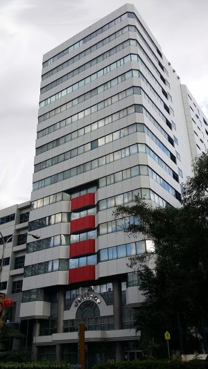 The Jollibee Centre, as seen from the sidewalk on the opposite side of San Miguel Avenue. The building is blocky in shape and has rows of concrete walls and glass windows.