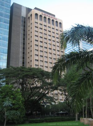 Lepanto features a thin cuboid shape in front of a larger rectangular prism. The smaller cuboid has beige color, horizontal recesses with glass windows, and semi-oblong-shaped cavities on top. The bigger cuboid has a dark brown color with two columns of glass panels. Shown here is Lepanto, as seen from somewhere near the middle of Ayala Triangle Park.