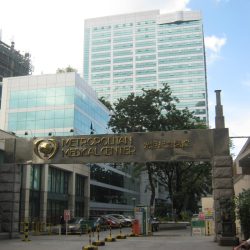 Metropolitan Medical Center is a high-rise cuboid building with a gray and blue-green facade of tiles and glass windows. This picture was shot from the sidewalk just across its entrance.