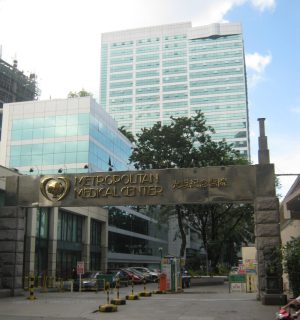 Metropolitan Medical Center is a high-rise cuboid building with a gray and blue-green facade of tiles and glass windows. This picture was shot from the sidewalk just across its entrance.