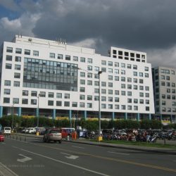 One E-com Center is one low-rise building that looks like two separate buildings, a white rectangular one and a gray square; both have concrete exteriors and several small square windows. This is One E-com Center as seen from Palm Coast Dr. cor. Seaside Blvd.
