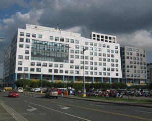 One E-com Center is one low-rise building that looks like two separate buildings, a white rectangular one and a gray square; both have concrete exteriors and several small square windows. This is One E-com Center as seen from Palm Coast Dr. cor. Seaside Blvd.