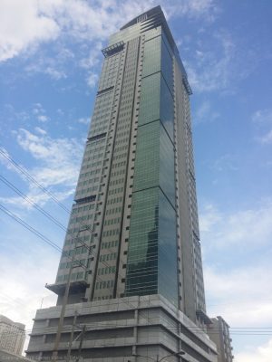 One San Miguel Building is a tall vertical structure with a facade of concrete and glass panels. This picture was taken from the opposite corner at Sheridan St. cor. Shaw Boulevard.