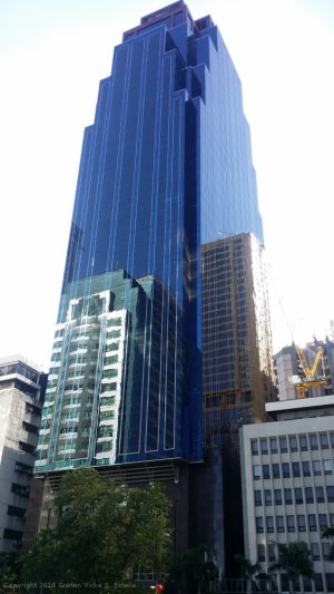 The Orient Square is a tall cuboid building with a facade of blue glass panels and maroon marble tiles. This is Orient Square, as seen from the parking between Emeral Avenue and Garnet Road near Julia Vargas Ave.