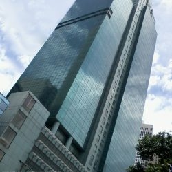 Petron Megaplaza, a modern glass skyscraper reflecting the surrounding cityscape under a cloudy sky shot from front of ECC (Employment Compensation Commission) Building. The partially visible building on the upper-left is BPI Buendia Center.