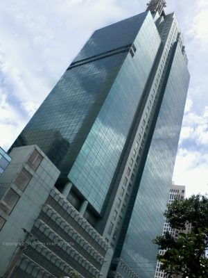 Petron Megaplaza, a modern glass skyscraper reflecting the surrounding cityscape under a cloudy sky shot from front of ECC (Employment Compensation Commission) Building. The partially visible building on the upper-left is BPI Buendia Center.