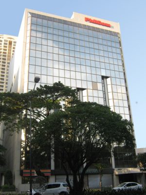 The low-rise building is almost square and has an exterior made of glass panels. This is the view from the sidewalk on the other side of Paseo De Roxas, indirectly across it (directly across the Paseo Center).