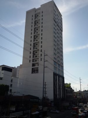 The Privato Ortigas Hotel is a high-rise building with small square glass windows on its upper 2/3. Its lower 1/3 with a larger area has a branching plant-like design in front, which seems to be made of metal. The side of the building visible in this photo has recesses at every floor on the upper 2/3 containing what appears to be external parts of airconditioning units. The side of the lower 1/3 is a flat concrete wall as if it's a firewall.