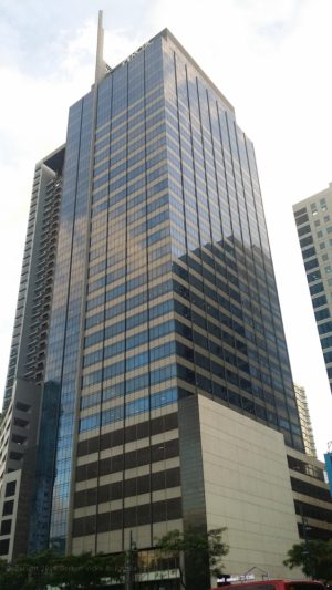 A.T. Yuchengco Centre is a tall square prism covered with beige and blue glass panels. Its lower floors have an exterior of a gray concrete podium. The picture was taken from 5th Ave. cor 26th St. in front of Six Neo.