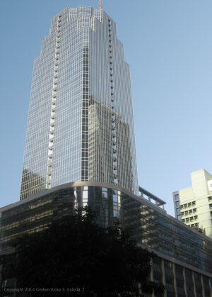 A tall skyscraper with a glass facade, set against a clear blue sky, with a wider podium structure and trees at its base.