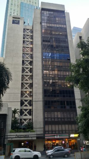 The Sagittarius Building is a gray mid-rise building. As seen in front, it has two parts. What appears to be the main section on the right is full of dark glass windows, with 7-11 and Subway at the ground floor.