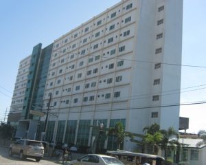 Santa Ana Hospital is a low-rise rectangular building with a facade of white concrete, green glass panels, and several small windows. This picture was taken from the sidewalk on the opposite side.