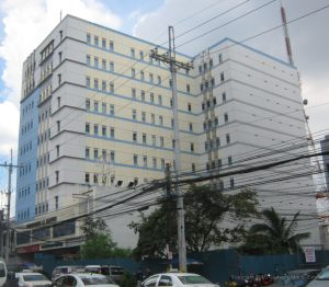 Sunnymede IT Center has a facade of mainly white concrete with small windows. Its structure has an L-shaped cross-sectional area. This is the view from the island dividing Quezon Ave. directly in front of the Triumph Building.
