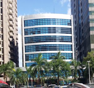 Suntree Tower is a cuboid structure atop a shorter cuboid. Its facade is that of gray tiles and blue glass panels. This is Suntree, seen from along Garnet Road with a parking space between it and Sapphire Road.