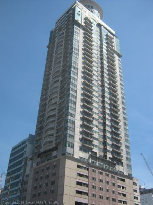 Sunview Palace is a tall rectangular prism with a cylindrical structure on top. Its colors are white and light maroon, and it has several windows and balconies. This is the view from the other side of Kalaw St., approximately where the opposite corner will be situated should Del Pilar St. extend beyond the intersection.