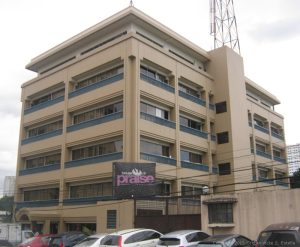 Sysu Centre standing as seen from the front of Cacao Hotel on the opposite side of Sgt. Esguerra St. Sysu Centre is a beige blocky low rise building with dark gray railings and glass windows from the second to the fifth floor. The ground floor has larger glass windows flanking the main entrance. The top floor has smallar area and recessed inward as if it's a penthouse. OPn the right side is a rectangular shaft that runs from ground to top floor, probably an elevator shaft.