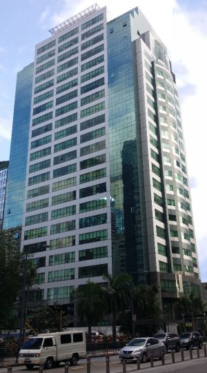 The Taipan Place is a tall and narrow cuboid building with a facade of blue glass panels, gray tiles, and dark marble. This is The Taipan Place, as seen from the sidewalk on the opposite side of Emerald Avenue.