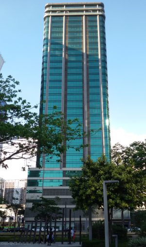 The upper floors of The Trade and Financial Tower have a rectangular-shape overall structure. From the Track 30th, where this shot was taken, the side that can be seen here has a facade made of green glass windows divided by vertical maroon and white segment. The top two floors have white horizontal protruding segments above, below, and between them. The lower levels with the wider area also have white horizontal segments between them. The top two among these floors have the same green glass windows, the ground floor has transparent glass walls, and the levels between them have walls made of maroon concrete.