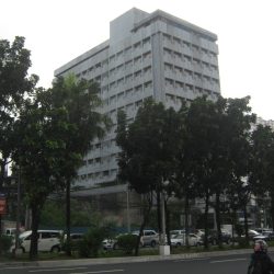 The building features a straightforward cuboid structure with a gray concrete exterior and evenly spaced windows. The shot was taken from the sidewalk on the southbound side of Quezon Ave.