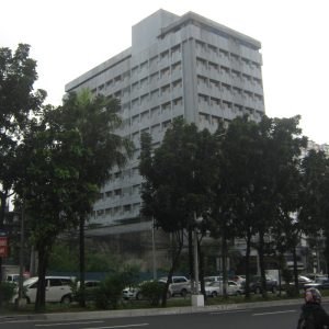 The building features a straightforward cuboid structure with a gray concrete exterior and evenly spaced windows. The shot was taken from the sidewalk on the southbound side of Quezon Ave.