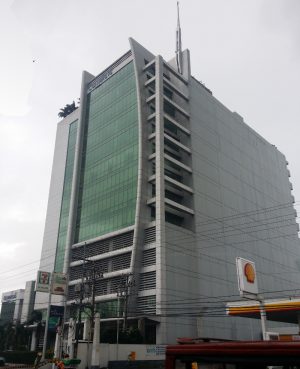 Two Sanparq as seen from across the Shell gasoline station adjacent to it. It has a large sign that says “Two Sanparq” on the top floor. The image also shows some cars and motorcycles parked in front of the tower, as well as some trees and plants on the sidewalk. The image was taken from a low angle, giving a sense of height and perspective to the tower.