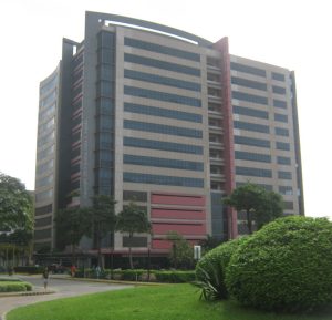 Vertex One's cube-shaped structure showcases predominantly black, red, and gray colors with dark glass window panels. This picture is as seen from the roundabout in front of the gate of Celadon Park.